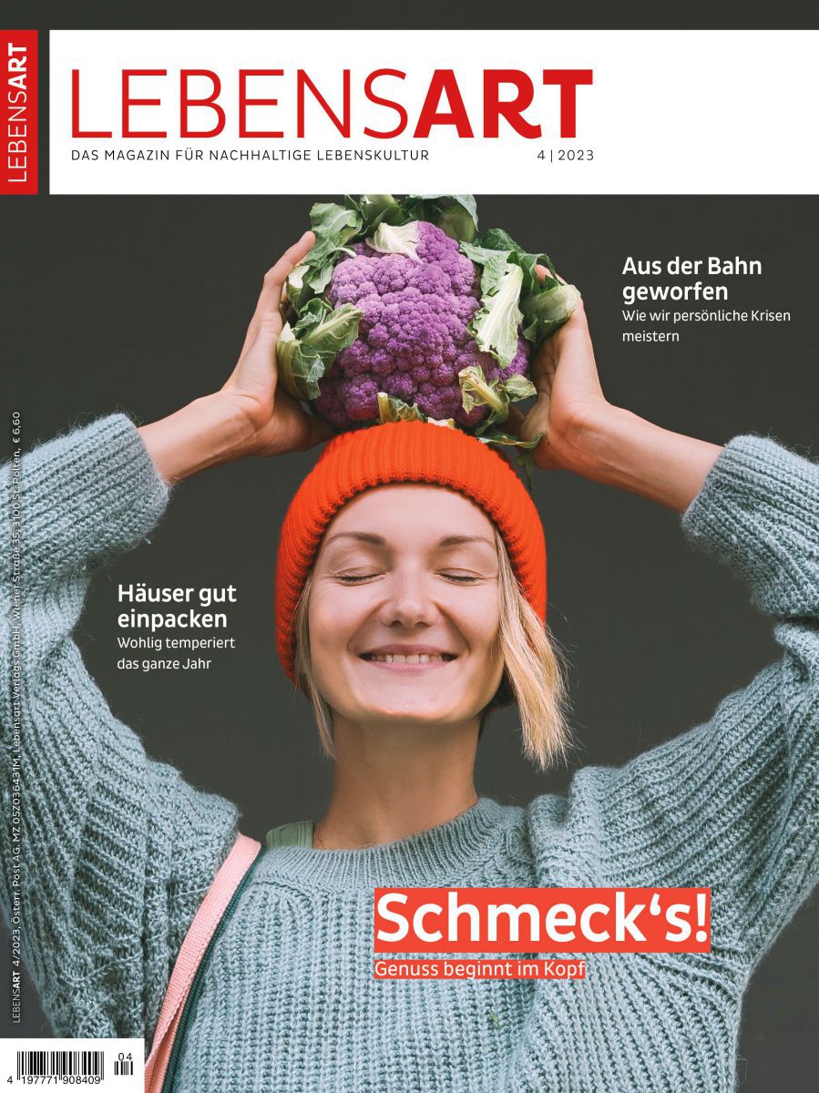 Eine Frau mit oranger Haube und grauem Pullover steht mit geschlossenen Augen und stolzem Lächeln vor dunkelgrauem Hintergrund und trägt eine violetten Karfiol auf ihrem Kopf.